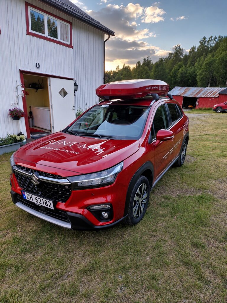 Roter Leihwagen mit Dachbox vor einem Bauernhaus im Sonnenuntergang